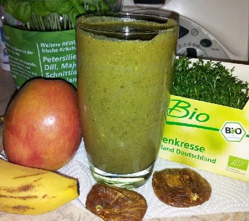 Glas mit grünem Smoothie, Zutaten wie Apfel, Banane und Kresse darum verteilt